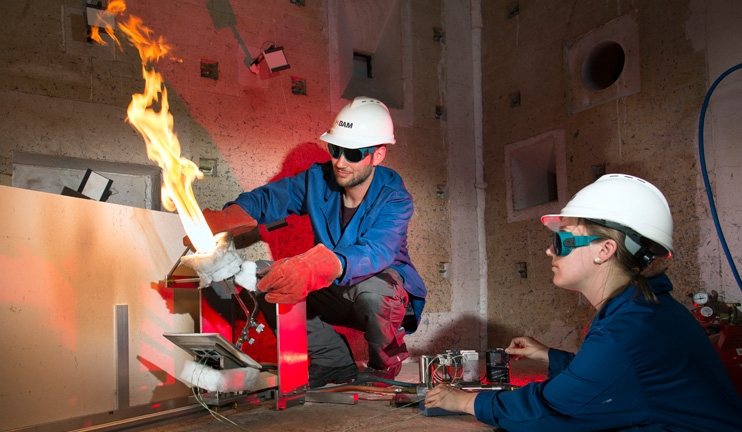Two BAM employees direct a burner flame onto a sample. Two BAM employees direct a burner flame onto a sample.