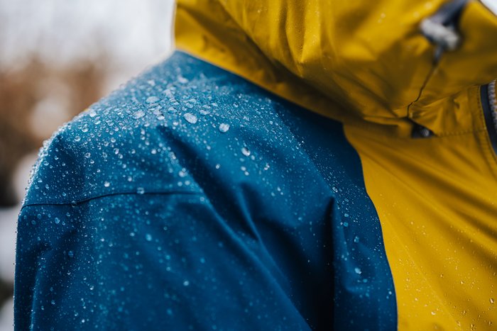 Nahaufnahme einer blau-gelben Outdoor-Funktionsjacke mit abperlenden Regentropfen.