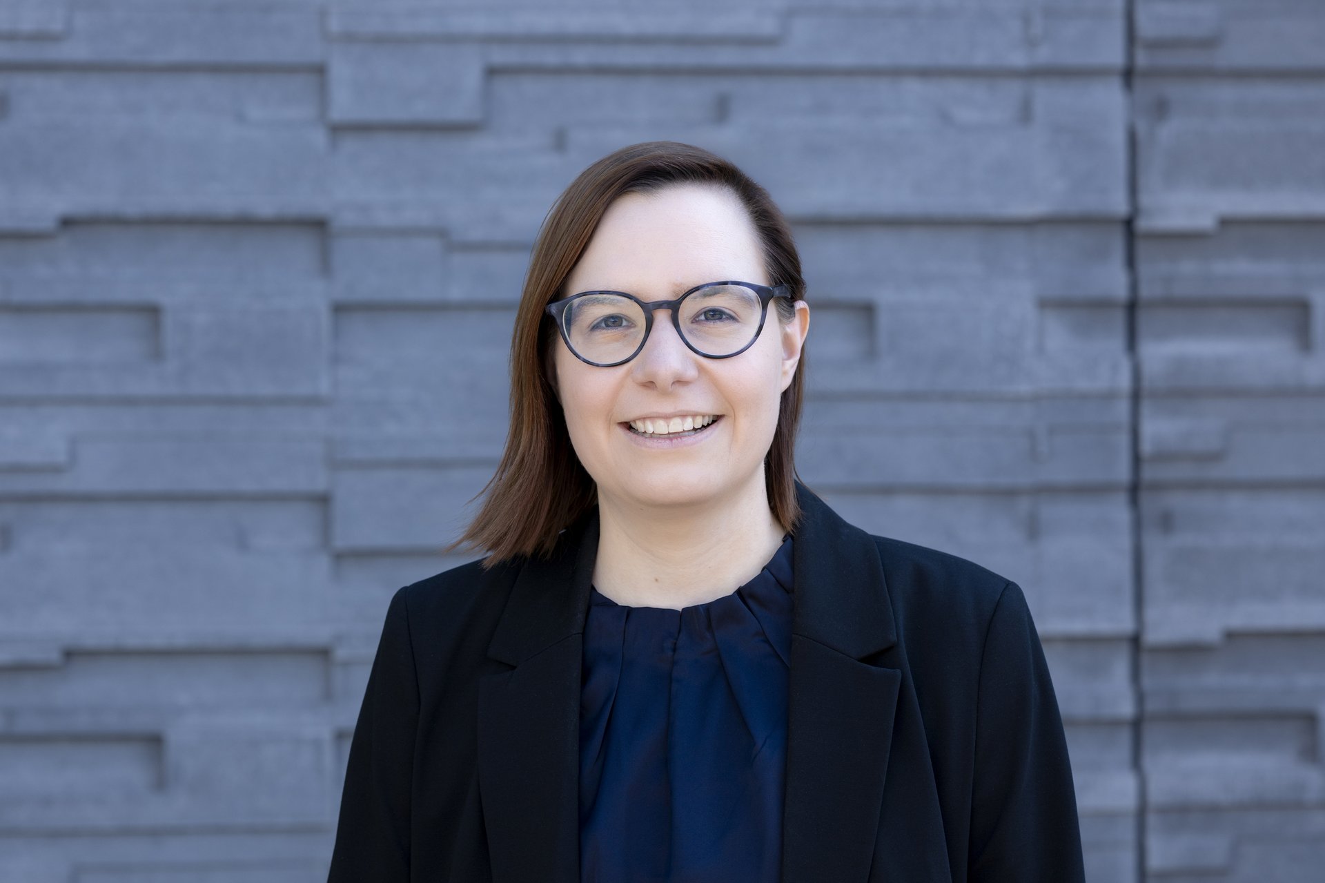 Janine George Wissenschaftslerin Janine George vor grauem Hintergrund in schwarzem Blazer, schwarz umrandeter Brille